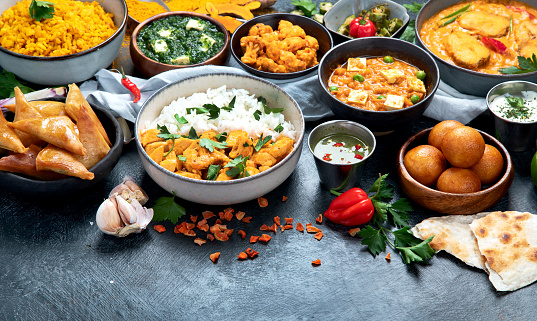 Dishes of indian cuisine. Curry, butter chicken, rice, lentils, paneer, samosa, spices. Bowls and plates with indian foodon dark on dark background. Top view with copy space.