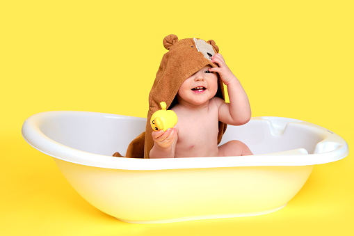 Happy baby toddler boy with hooded towels in a white tub on a studio yellow background. A smiling child at the age of one year, copy space