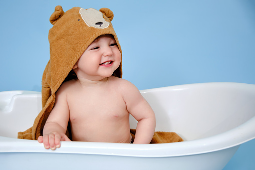 Happy baby toddler boy with hooded towel in a white bathtub on a studio blue background. A smiling child at the age of one year