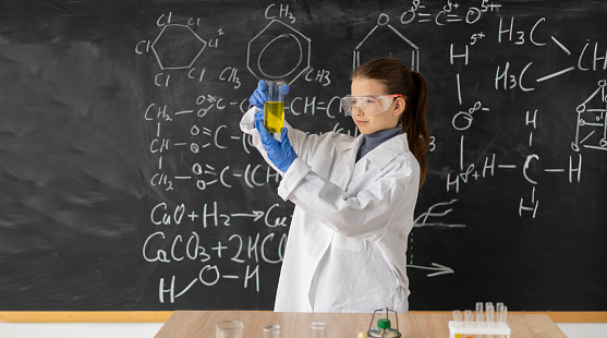 A child genius is wearing a bow tie and a lab coat while conducting scientific experiments in a classroom. She is wearing protective goggles and smiling while looking at the camera.
