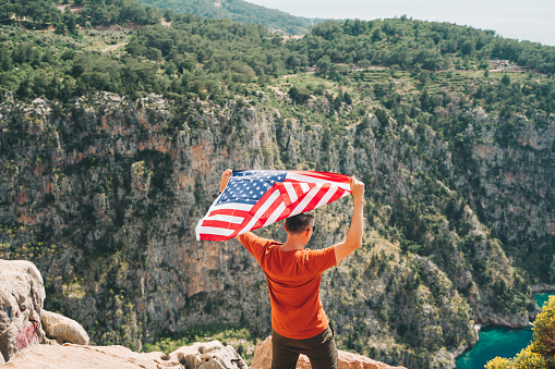 Young man standing on a rock cliff and waving the US flag while looking at sea beneath. Veteran traveller waving the American flag while standing on a mountain top. 4 July Independence Day