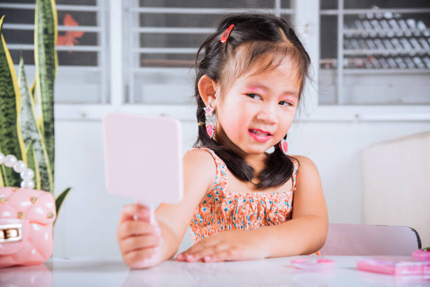 Asian adorable funny little girl making makeup her face and looking face herself in the mirror, Learning activity to be woman, happy kid is beautiful make up with cosmetics toy