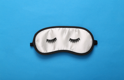 Sleep mask with closed eyes and eyelashes on blue background. Top view, flat lay. Concept of vivid dreams. Minimal style