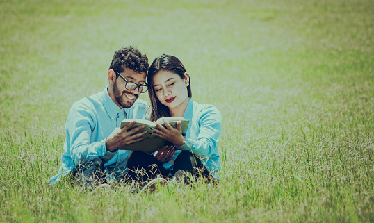 Two people sitting on the grass reading a book, concept of people reading outside, A couple sitting on the grass reading a book