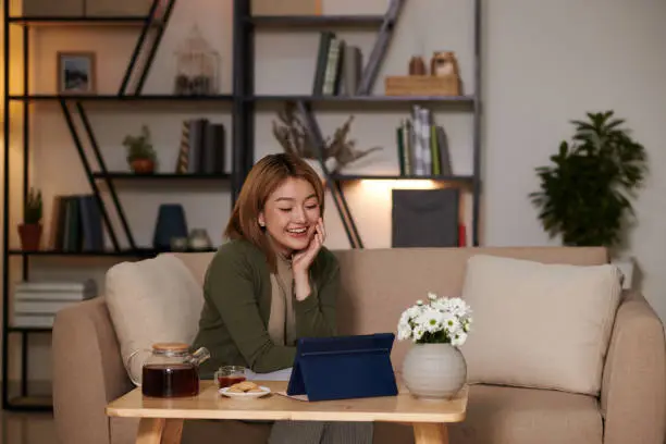 Cheerful young woman having tea and homemade cookies when watching new episode of romcom on tablet computer