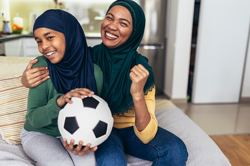 Football match watching concept. Muslim woman and young daughter sit couch cozy celebrate soccer team victory.