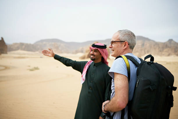 Waist-up view of smiling Middle Eastern man in traditional attire gesturing to off-camera landmark as he and mature man with backpack explore Al-Ula.