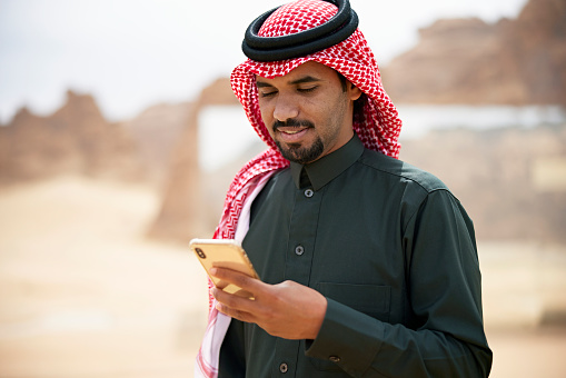 Waist-up view of Middle Eastern man with mustache and goatee in black dish dash, kaffiyeh, and agal checking mobile device.