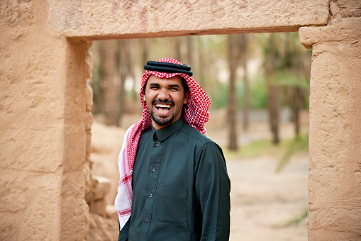 Waist-up view of laughing Middle Eastern man in traditional attire standing under mudbrick archway and looking at camera.