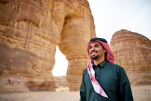 Waist-up view of man in black dish dash, kaffiyeh, and agal looking away from camera and smiling with famous Elephant Rock monolith in background.