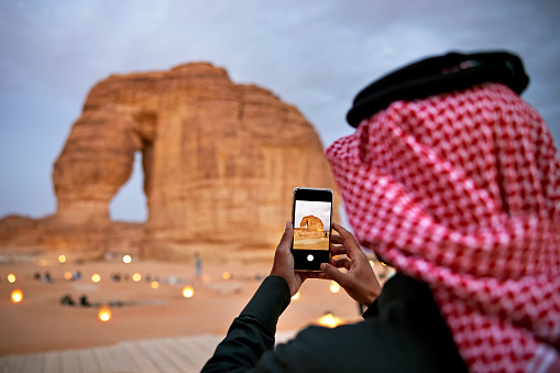 Over the shoulder view with focus on device as Middle Eastern man in traditional attire captures a memory on mobile device of Al-Ula landmark.