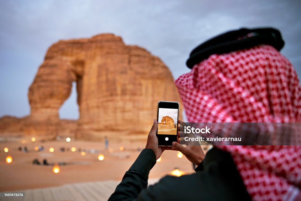 Saudi man photographing Elephant Rock with smart phone Over the shoulder view with focus on device as Middle Eastern man in traditional attire captures a memory on mobile device of Al-Ula landmark. Middle East Stock Photo