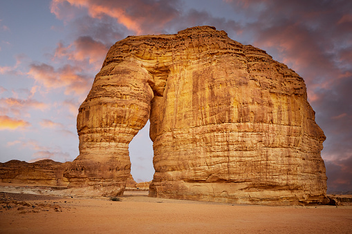 Full view of red sandstone monolith shaped by wind and rain erosion over millions of years against background of blue sky and pink clouds at sunset.