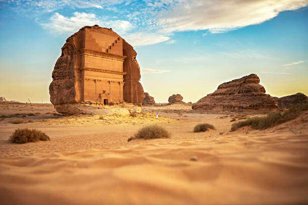 Distant view of Saudi man exploring Hegra in Medina Region Surface level view from sand dune with focus on background Tomb of Lihyan, son of Kuza, iconic burial chamber cut into existing rock formation. arabian peninsula stock pictures, royalty-free photos & images