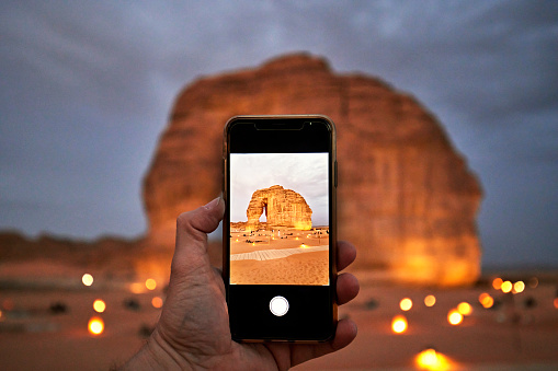 Focus on foreground hand holding mobile device vertically while photographing local landmark at dusk, a monolith eroded by wind and rain for millions of years.