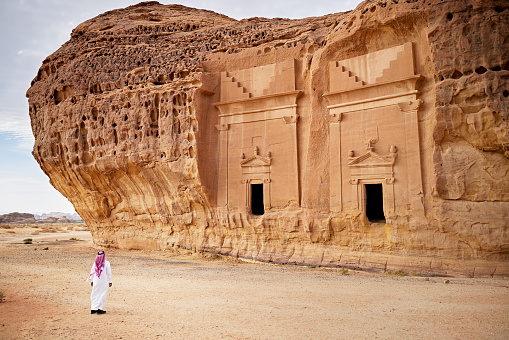 Full length view of Middle Eastern man in traditional attire visiting ancient Jabar Al Ahmar burial chambers cut into existing rock formations.