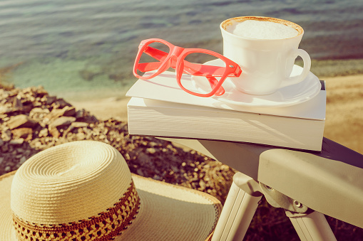 Reading on holidays. Coffee cup and book on chair outdoors against blue sea water background.