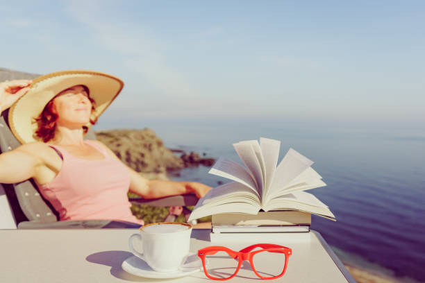 Reading on holidays. Mature tourist woman relaxing on coast, read book, enjoy sea view from cliff. Focus on coffee cup and glasses.