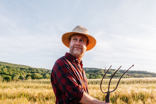 Color image depicting a mid-adult farmer standing in a wheat field surveying his crops. He is wearing a straw hat and red and navy blue check shirt.