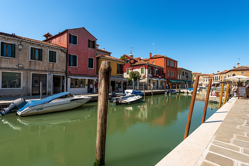 Murano, Italy - June 2th, 2021: Small sea channel with moored boats in Murano island, Venice lagoon, UNESCO world heritage site, Veneto, Italy. Europe. This island is famous for the production of artistic glass. A group of tourists and locals stroll along the narrow street that borders the canal on a sunny spring day with clear sky.