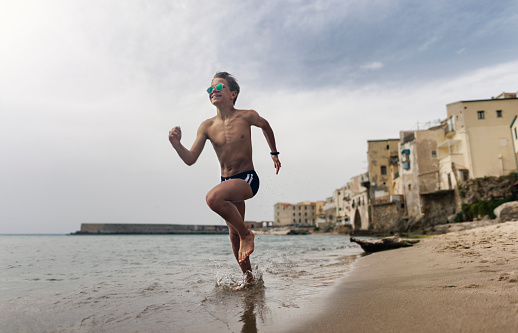 Teenage boy is playing on small, old town beach. Vacations day in Cefalu, Sicily, Italy.\nShot with Canon R5