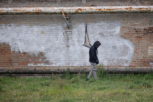 Parma, Italy - march 2022: Boy with a Black Hoodie in his Head Walking on the Grass Next to a Brick City Wall.