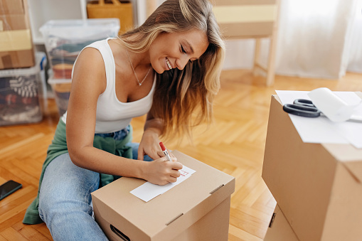 Young woman sitting on the living floor and labelling cardboard boxes. She is preparing for moving