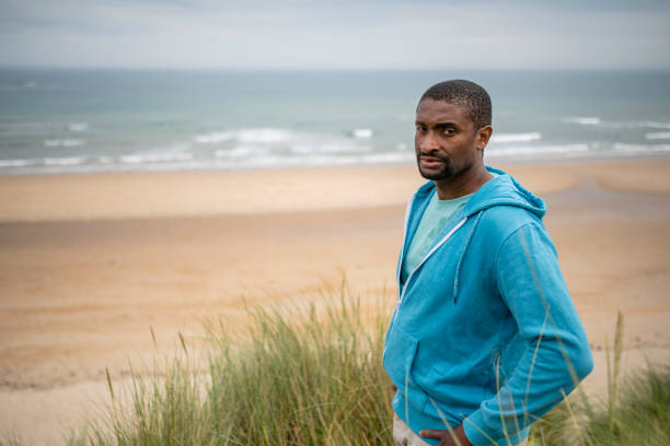 A mid adult man standing in the grasses in a sand dune at Beadnell beach, North East England. He has his hands on his hips and is looking at the camera with a serious look on his face.