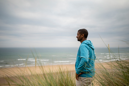 A mid adult man standing in the grasses in a sand dune at Beadnell beach, North East England. He has his hands on his hips and is looking at the view.