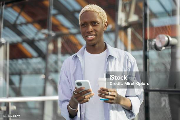 Smiling African American hipster using mobile phone holding cup of coffee standing on the street