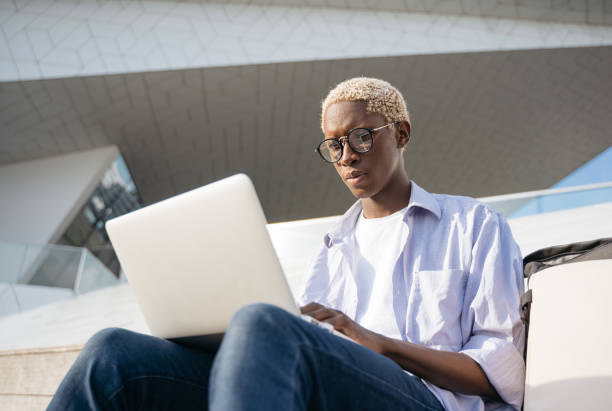 African American programmer using laptop computer working freelance project sitting at workplace. Pensive student wearing stylish eyeglasses studying sitting in university campus stock photo