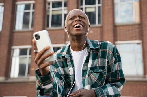 Authentic portrait of emotional  hipster guy with stylish hairstyle using mobile phone listening music, laughing outdoors. Happy African American man holding smartphone communication on the street