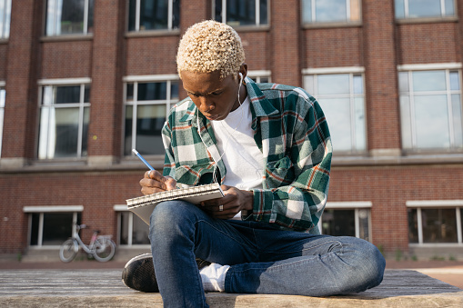 Portrait of pensive man artist wearing casual clothing drawing  something sitting alone on the street. Young stylish writer taking notes holding notepad, listening music, relaxing outdoors