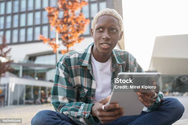 Smiling African American student using digital tablet studying, learning language sitting in university campus, online education concept. Stylish freelancer working outdoors