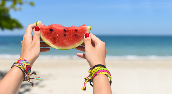 Womens hands with friendship bracelets holding watermelon against beach
