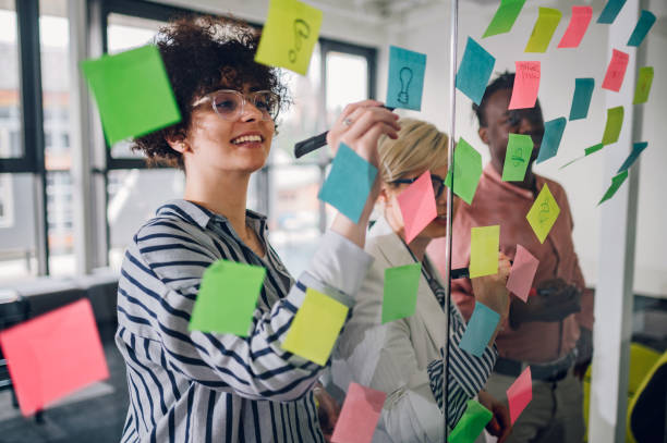 team multirazziale al lavoro che scrive idee su note adesive sulla parete di vetro - spremersi le meningi foto e immagini stock