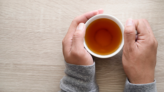 First person top view photo of male hands in grey sweater touching a white cup of tea on a vintage wooden table background with copy space