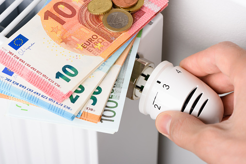 Hand adjusting the valve knob of heating radiator temperature thermostat with stack of euro money banknotes on it. Expensive heating costs and rising energy bill prices for winter cold season