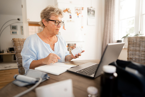 Senior woman is sitting by her kitchen table, engaged in an online consultation with her doctor or nurse. A so called telemedicine video call on her laptop at home.