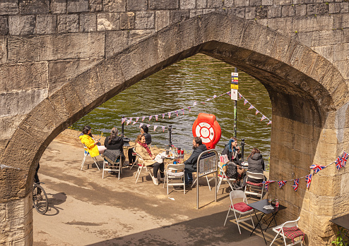 York, UK. May 31, 2022.  Outdoor cafe under a bridge and beside a river. People sit at tables and bunting decorates the scene.
