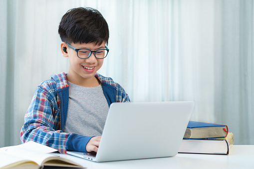 Happy handsome Asian preteen boy with blue jacket and eyeglasses smiling, videocalling, talking with friends in social media and learning online class with laptop computer in bedroom at home