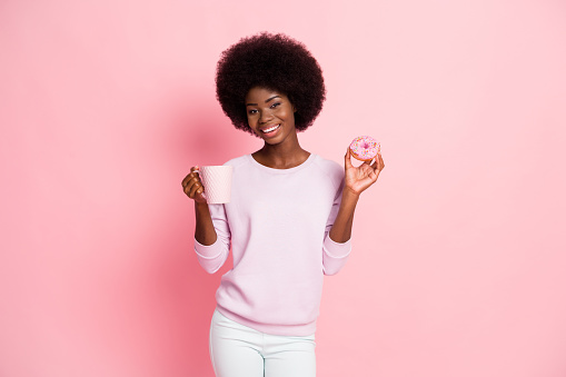Portrait of lovely cheerful wavy-haired girl drinking enjoying coffee latte eating sugary donut isolated over pink color background