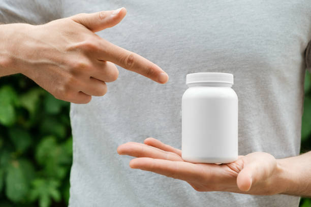 istock Man is holding white plastic jar in one hand and pointing to it with other against blurred green background. Close up, copy space. 1401509850