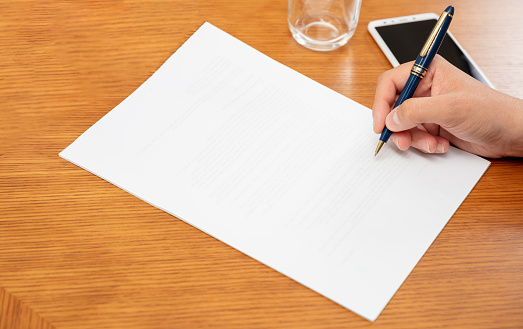 Man holding a pen on a blank white paper, close up above view, wooden office table background. Male hand signing on empty document, copy space