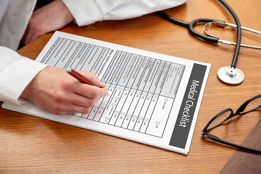 Medical checklist. Woman doctor holding a pen, close up view, wooden office table background. Female hand filling in the document, copy space