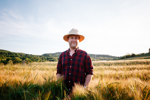 Color image depicting a mid-adult farmer standing in a wheat field. He is wearing a straw hat and red and navy blue check shirt. He is smiling, happy and contented.