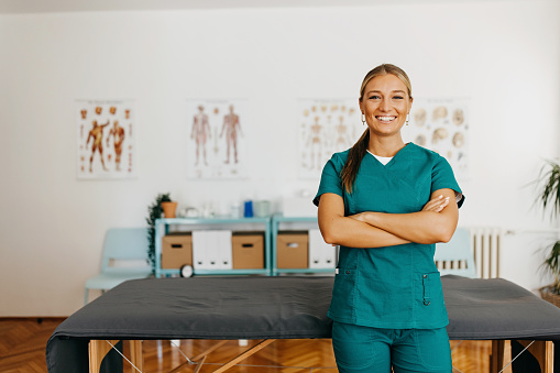 Beautiful blonde woman physiotherapist portrait standing next to medical table. Portrait of confident young female physiotherapist in green uniform,