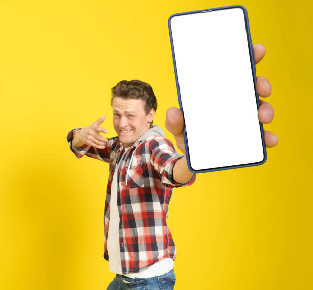 istock Happy to win handsome man pointing at smartphone with white empty screen, wearing red plaid shirt and jeans cellphone display mock up isolated on yellow background. Mobile app advertisement 1401515337