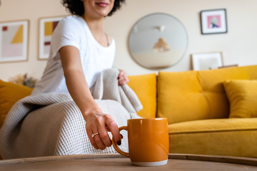 Unrecognizable woman hand grabs cup of coffee. Woman having tea sitting on the couch at home. Copy space. Lifestyle.