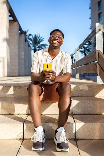 Happy, young African American man using mobile phone outdoors sitting on stairs. Vertical image. Copy space. Lifestyle concept.